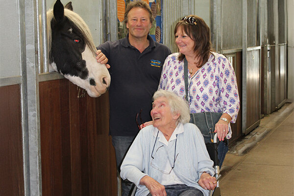 A horse stands in a stable with its head lowered over the door. A visitor is stroking the horses head, and beside them another visitor sits in a wheelchair.