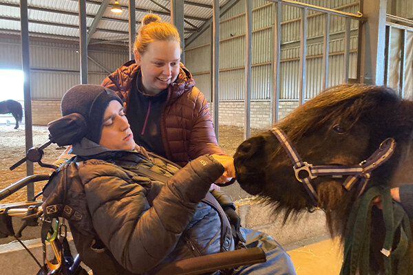 A wheelchair user is stroking the muzzle of a small, black pony, with the support of a carer.