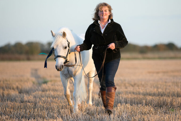 Woman leading grey horse through stubble field